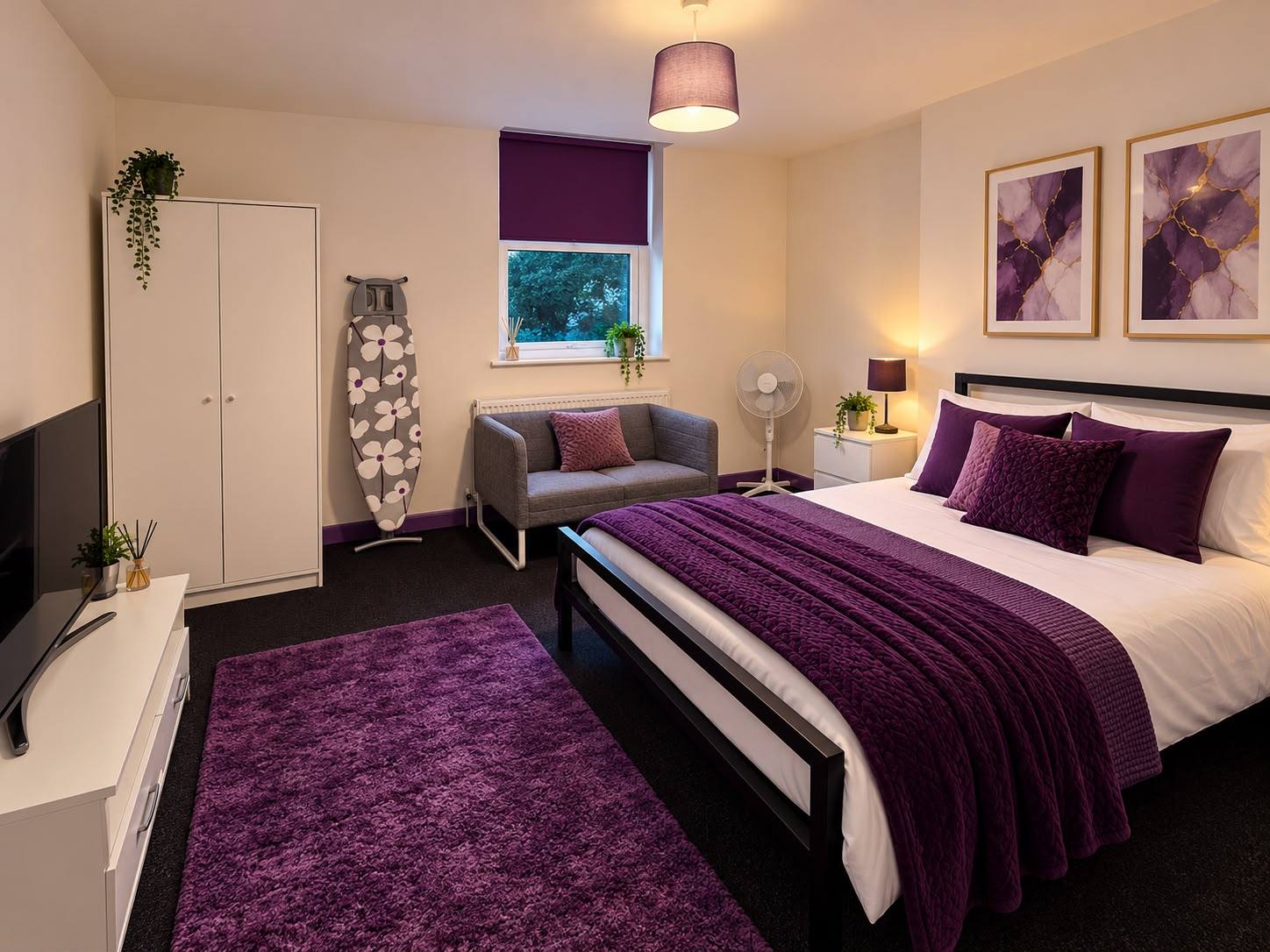A furnished bedroom with a double bed and purple throw, a white wardrobe, a small sofa, a TV unit and a rug.