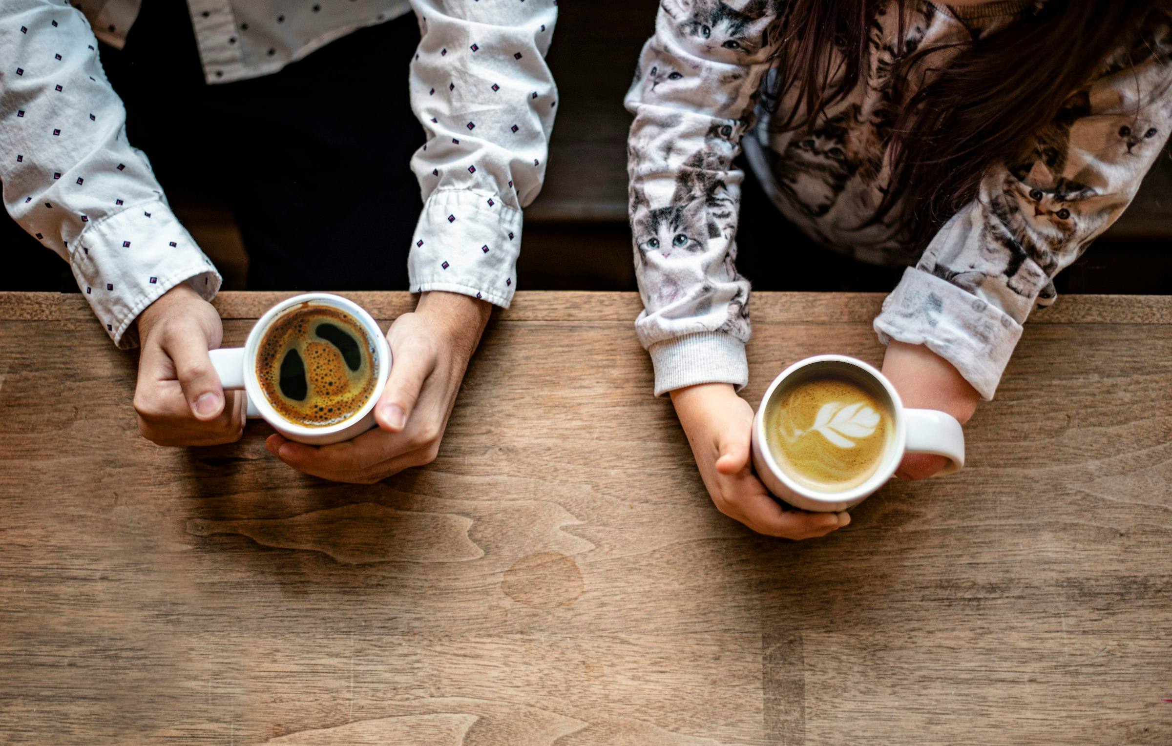 Two people sitting across a wooden table from each other, each holding a cup of coffee.